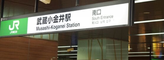 モダンな建物の武蔵小金井駅南口の看板。日本語（武蔵小金井）と英語訳が表示されています。近くには、個人向けの韓国語教室 (韓国語教室マンツーマン) を宣伝する小さなバナーがあります。.