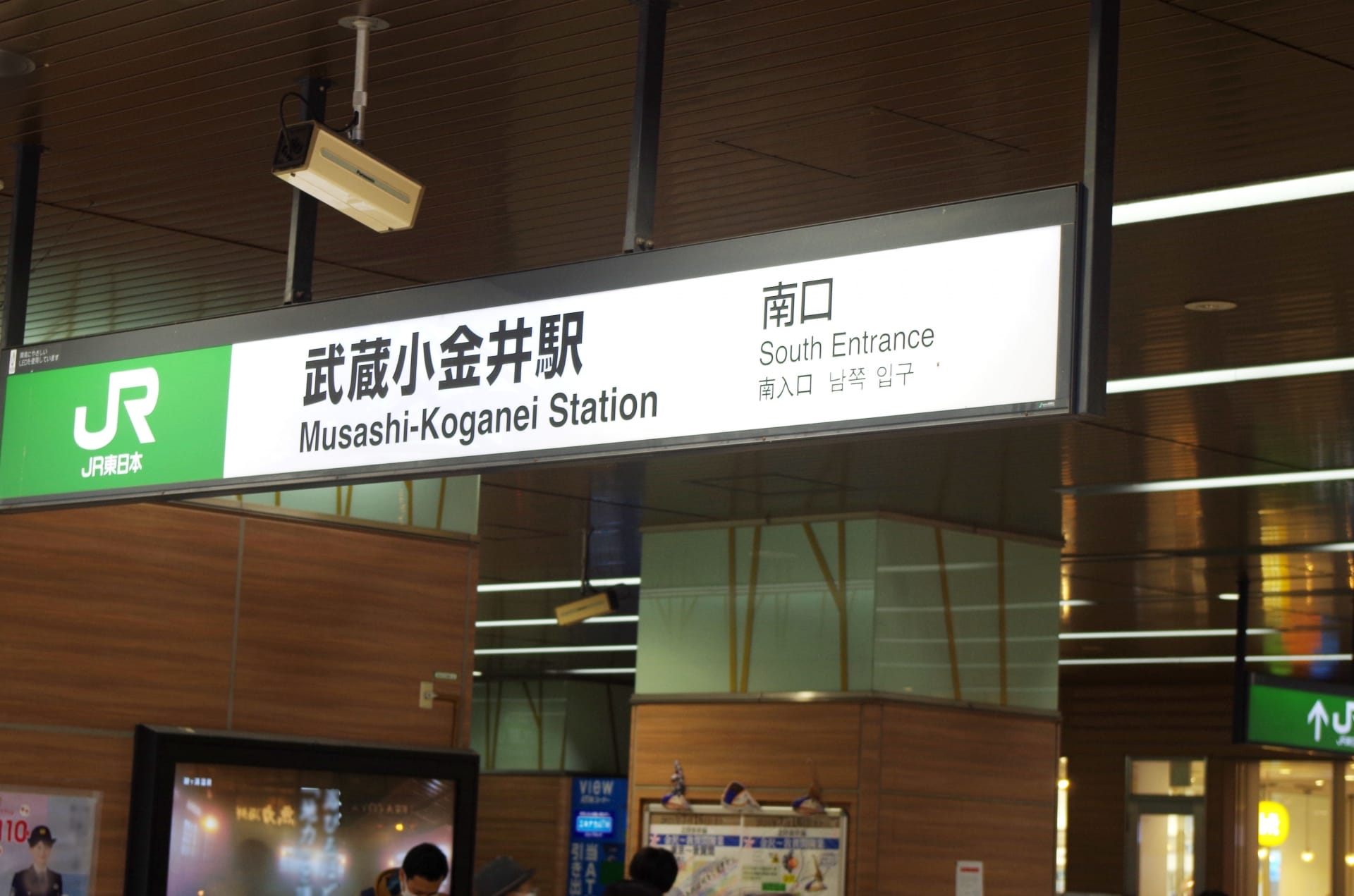 モダンな建物の武蔵小金井駅南口の看板。日本語（武蔵小金井）と英語訳が表示されています。近くには、個人向けの韓国語教室 (韓国語教室マンツーマン) を宣伝する小さなバナーがあります。.