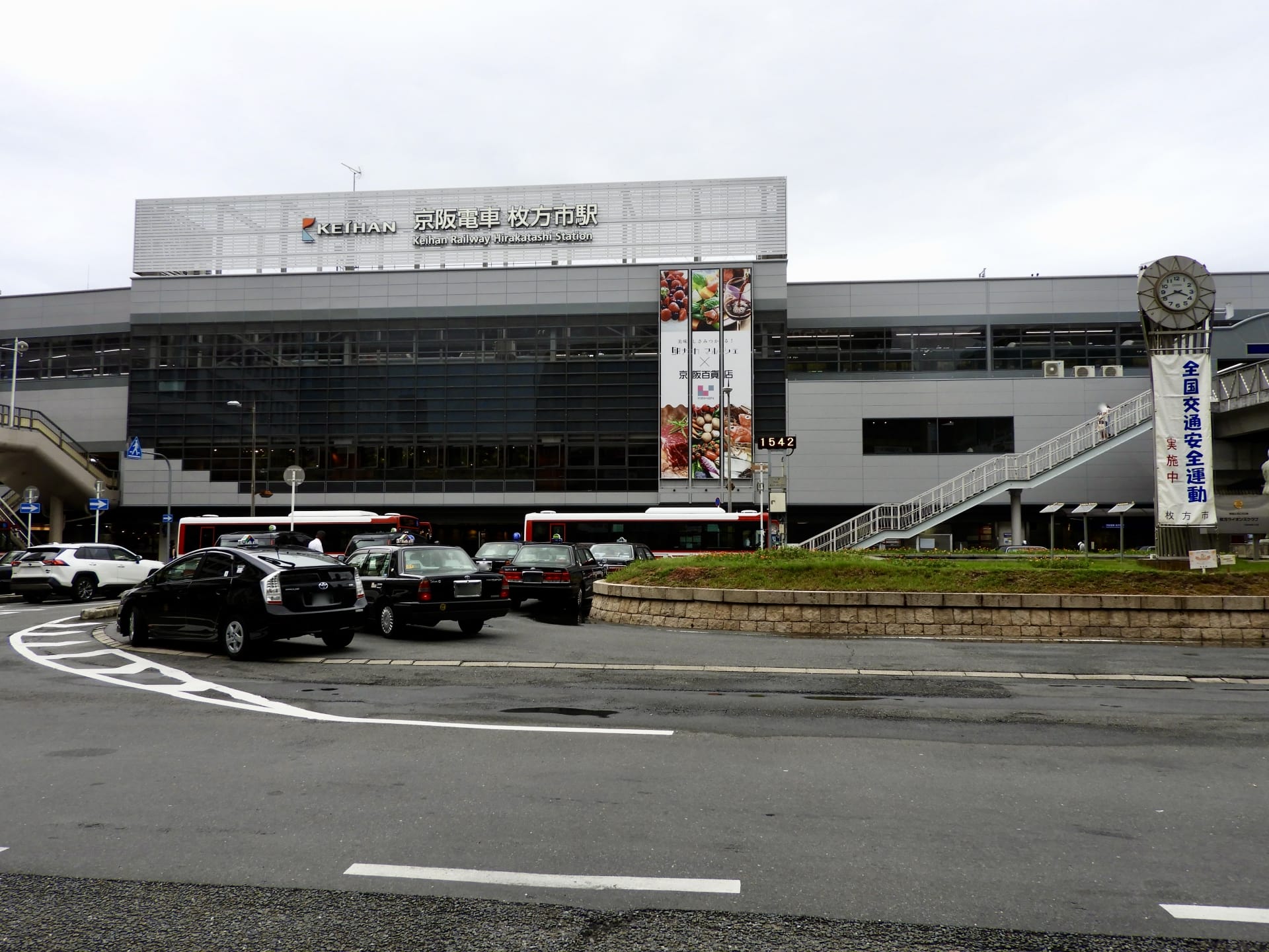 枚方市にある近代的な鉄道駅の外観。前をバスや車が通過し、ポールには時計があり、日本語と英語の文字が書かれた看板が目に見える。近くには韓国語教室マンツーマンがあり、マンツーマンで韓国語を学びたい方に最適です。.