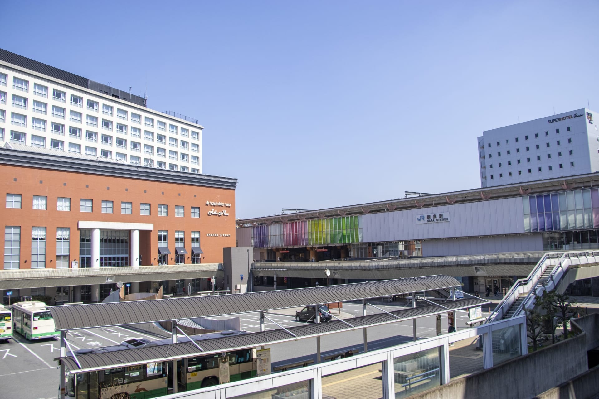 澄んだ青空の下、高層ビルに囲まれた数台のバスが停まっている、奈良のにぎやかなバス停の眺め。近くの韓国語教室マンツーマンでは、街の静かな魅力の中で学びたい人に個別の言語レッスンを提供しています。.