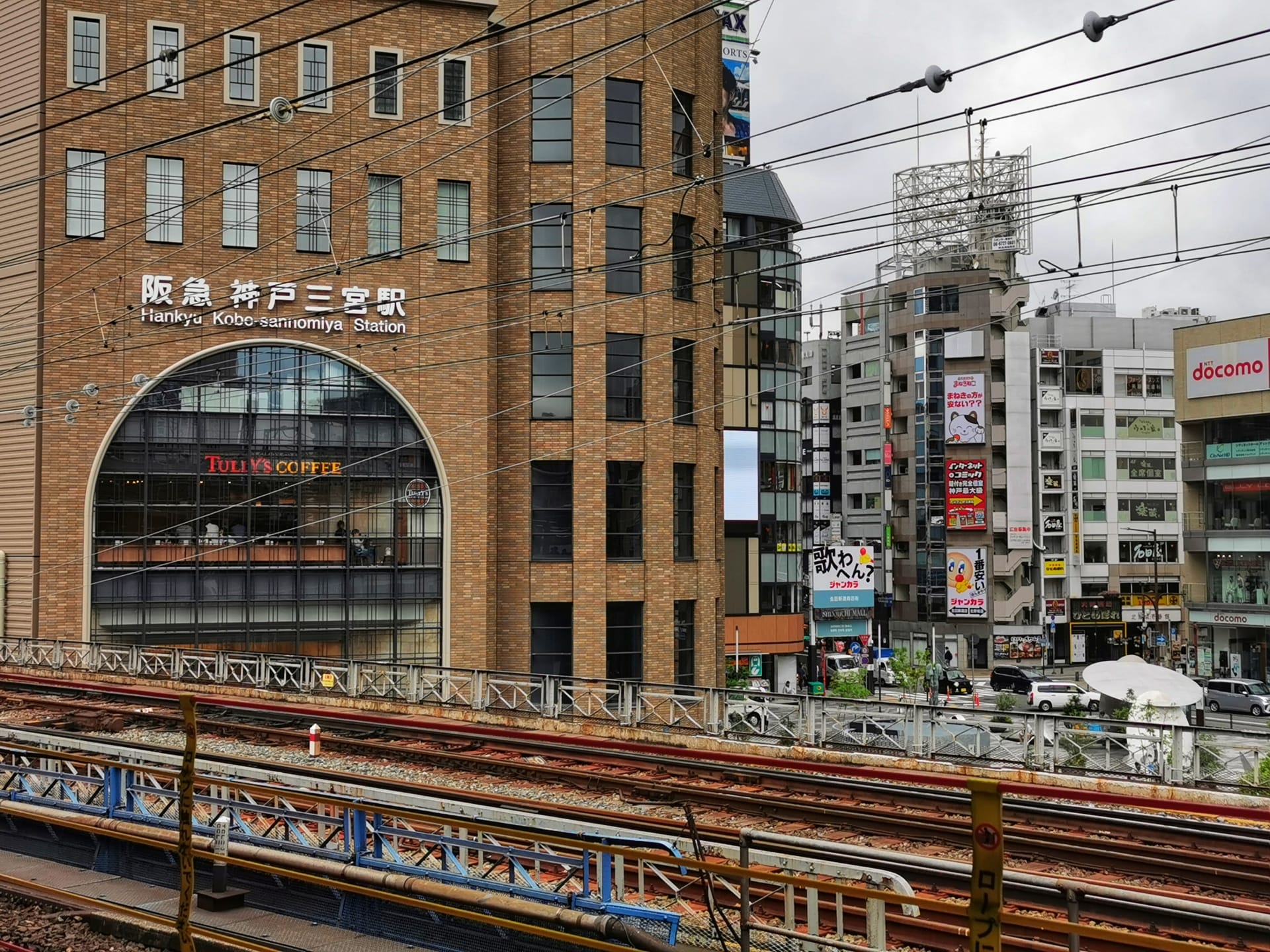 三宮にある阪急神戸三宮駅の駅舎を描いた都市風景。前景には複数の線路があり、さまざまな建物や看板が立ち並ぶ賑やかな街並みが広がっています。上には、活気に満ちた街の中心部に曇り空が広がっています。.