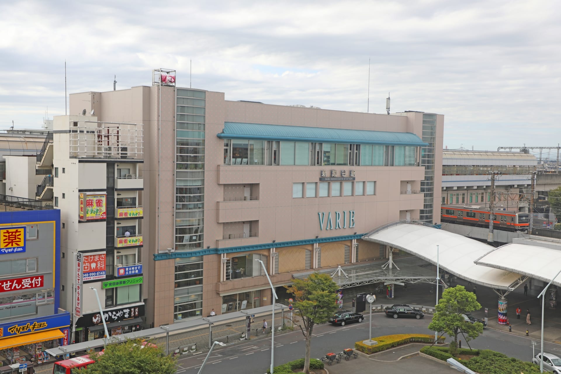 ガラス張りのファサードと高架を通過する電車が特徴的な高層ビル「VARIE」がある新越谷の都会的な風景。通りのレベルでは、いくつかの小さなお店や看板に囲まれた韓国語教室マンツーマンを見つけてください。.