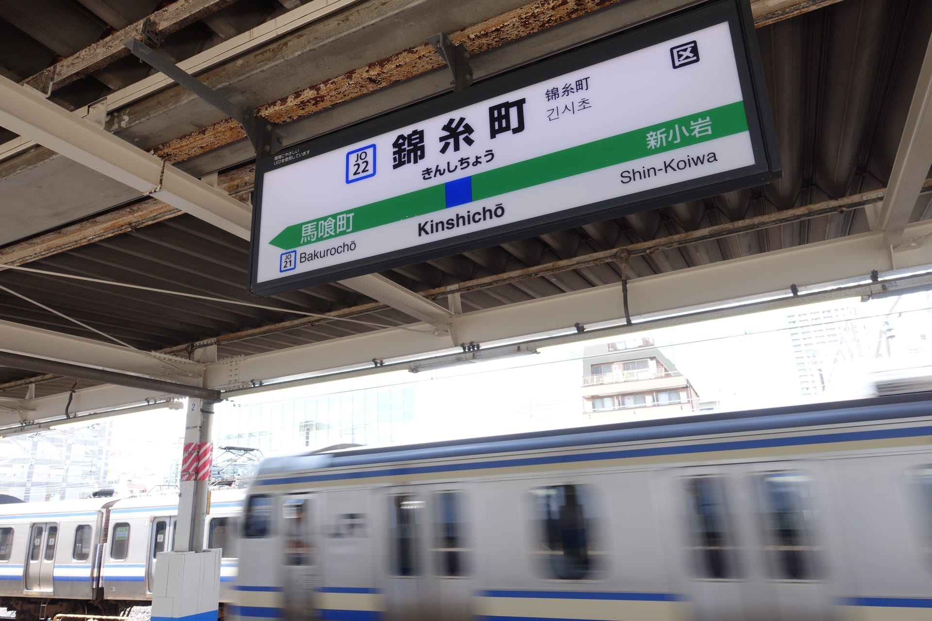 錦糸町駅のホームを電車が通り過ぎると、人気の錦糸町地区のすぐそばにある馬喰町と新小岩方面への案内標識が立っている。.