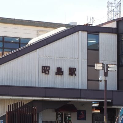 斜め屋根の建物の右側には日本語の文字と時計が掲げられています。建物には大きな窓があり、近くには昭島の韓国語教室マンツーマンの案内看板が立っています。.