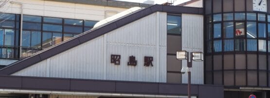 斜め屋根の建物の右側には日本語の文字と時計が掲げられています。建物には大きな窓があり、近くには昭島の韓国語教室マンツーマンの案内看板が立っています。.