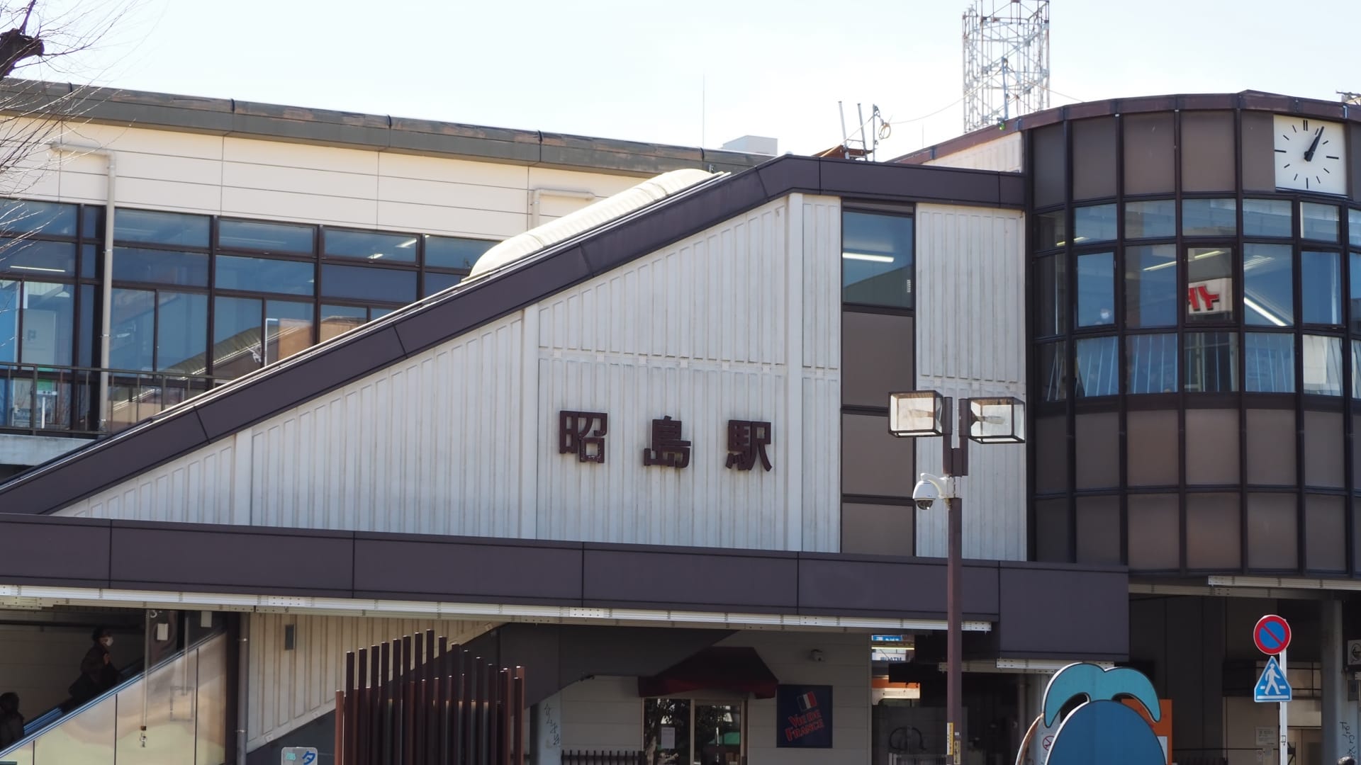 斜め屋根の建物の右側には日本語の文字と時計が掲げられています。建物には大きな窓があり、近くには昭島の韓国語教室マンツーマンの案内看板が立っています。.