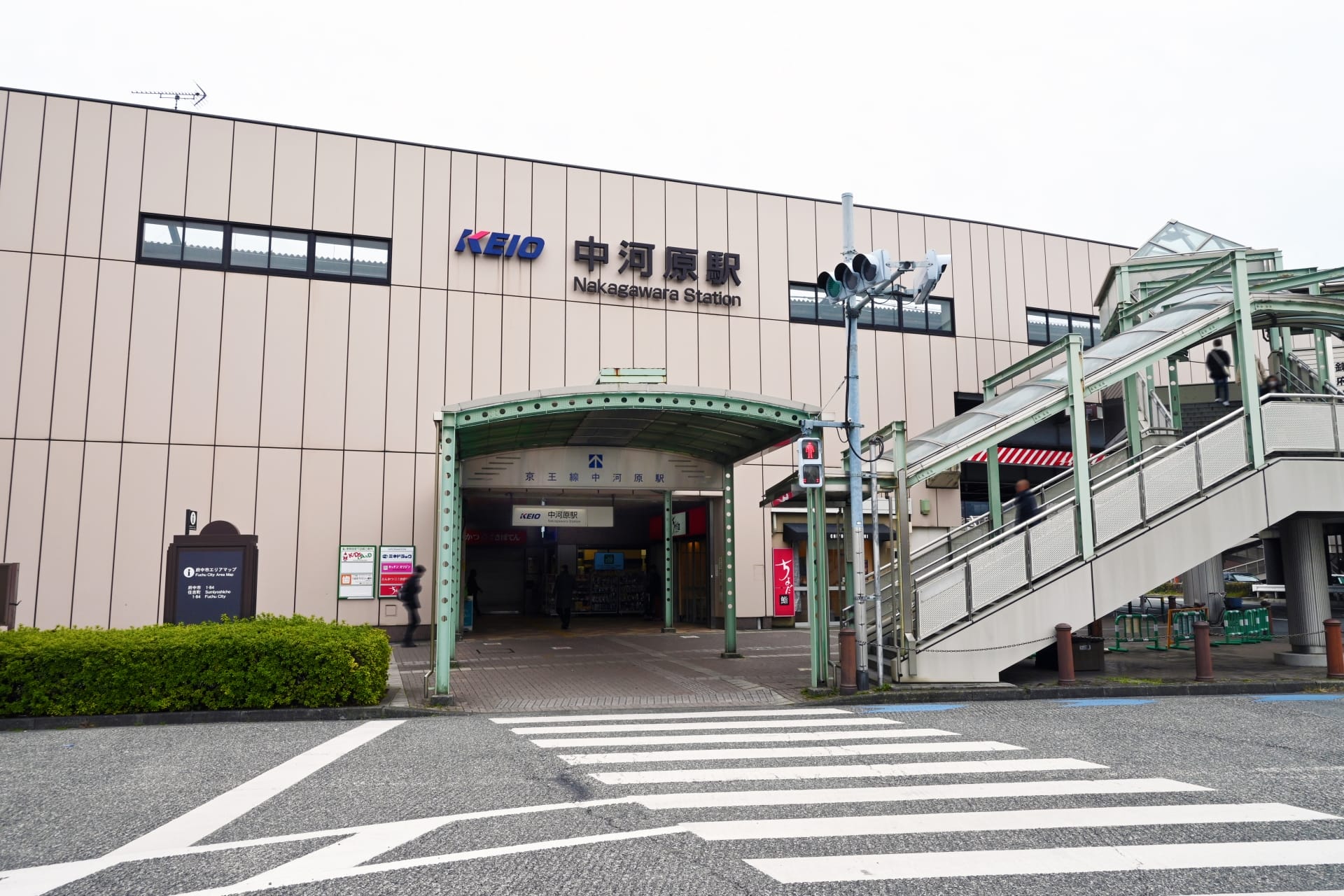 ベージュ色の外観と KEIO の看板が目印の中原駅の入り口には、手前に歩道と横断歩道へと続く階段があります。近くには、マンツーマンで学べる韓国語の個別クラスがあります。.