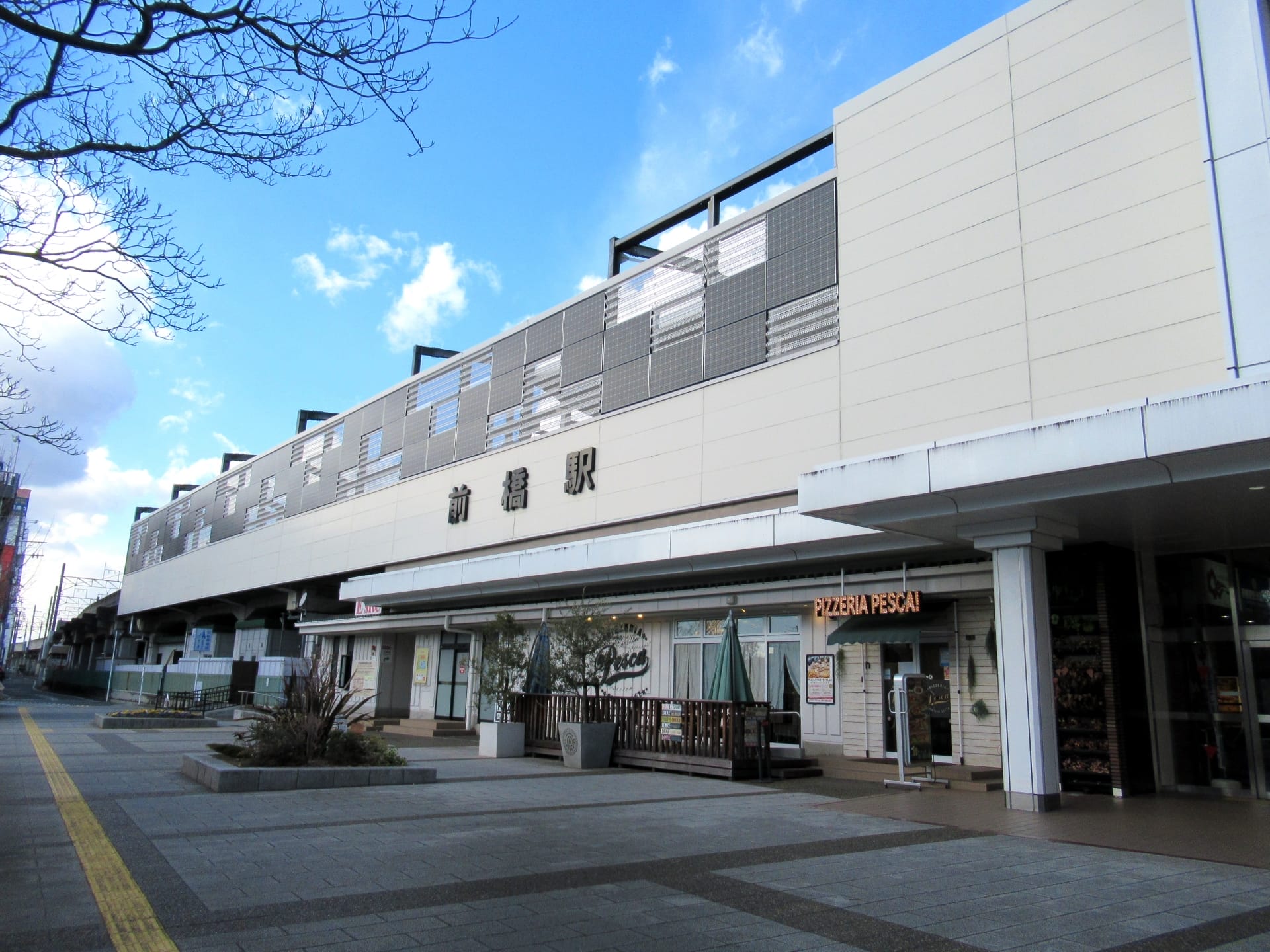 前橋・群馬の晴れた空の下にあるモダンな駅の外観。入口近くの居心地の良いピザ屋と活気に満ちたプランターが特徴。.