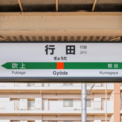 駅の標識には、都市のビル群を背景に、吹上、行田、熊谷方面への案内が書かれています。.