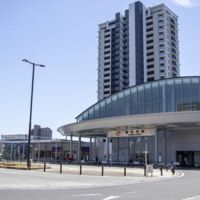 春日井の近代的な鉄道駅の入り口は、澄み切った青空の下の高層ビル群を背景に、洗練されたガラスのファサードと屋根付きの通路を誇っています。.