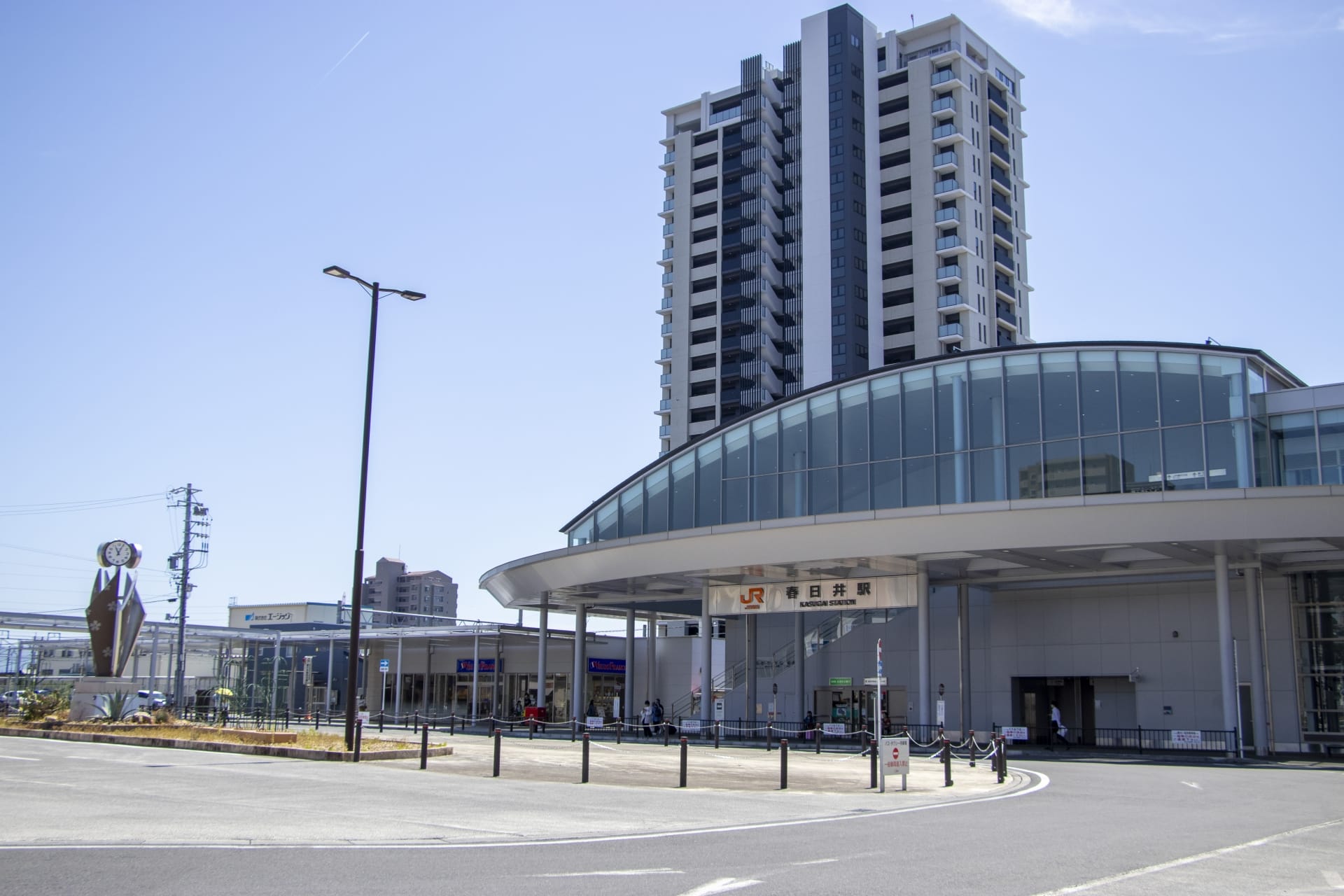春日井の近代的な鉄道駅の入り口は、澄み切った青空の下の高層ビル群を背景に、洗練されたガラスのファサードと屋根付きの通路を誇っています。. 春日井の近代的な鉄道駅の入り口は、澄み切った青空の下の高層ビル群を背景に、洗練されたガラスのファサードと屋根付きの通路を誇っています。.
