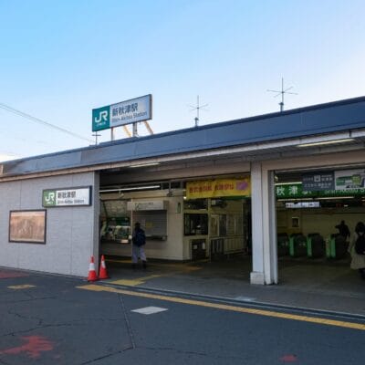 JR 西日暮里駅入口の外観。標識、改札口、駅に入る 2 人の人物。おそらく近くの韓国語教室マンツーマンに個別の言語レッスンを受ける途中だろう。.