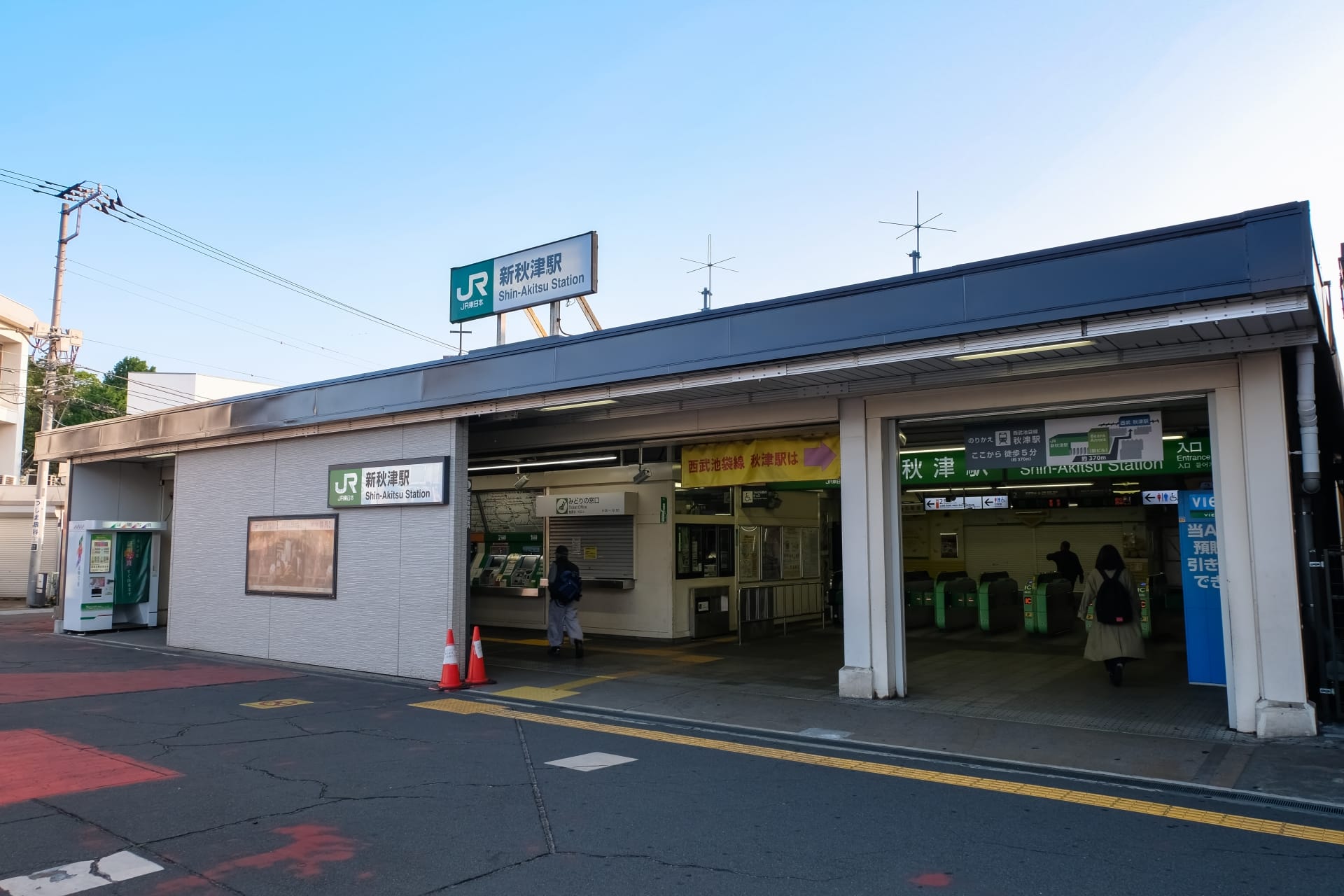 JR 西日暮里駅入口の外観。標識、改札口、駅に入る 2 人の人物。おそらく近くの韓国語教室マンツーマンに個別の言語レッスンを受ける途中だろう。.