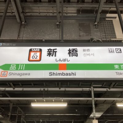 多言語で「新橋」を表示し、品川と東京への方向を示す駅看板。近くには、個人に合わせた韓国語レッスンを提供する韓国語教室マンツーマンがあります。.