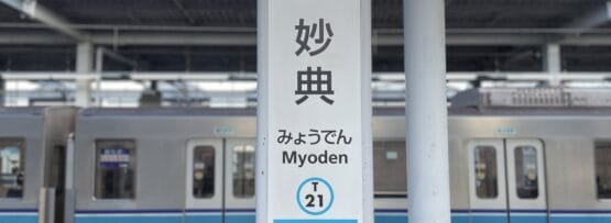 妙典駅の標識。背景には電車が写っており、駅名「妙典」が日本語、韓国語、英語で表示され、駅番号はT21となっている。.