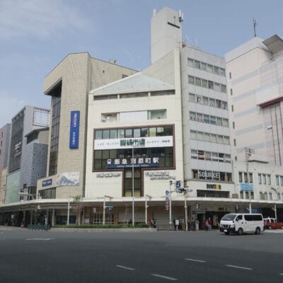 河原町の繁華街の交差点のストリートビュー。日本語の看板のある商業ビルが特徴。その中に韩国語教室マンツーマンがひっそりと佇んでいます。薄曇りの空の下、数台の車が道路を走っている。.