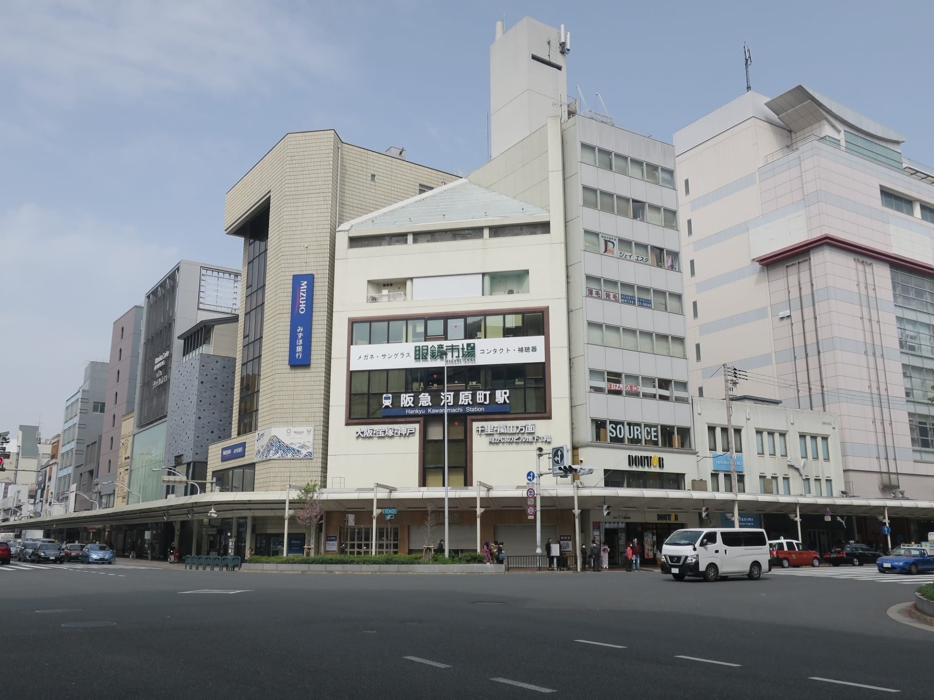 河原町の繁華街の交差点のストリートビュー。日本語の看板のある商業ビルが特徴。その中に韩国語教室マンツーマンがひっそりと佇んでいます。薄曇りの空の下、数台の車が道路を走っている。.