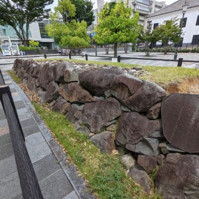 公園の舗装された歩道に沿って石積みの擁壁が並び、その背景には木々や建物が広がり、甲府の静かな雰囲気を醸し出しています。.