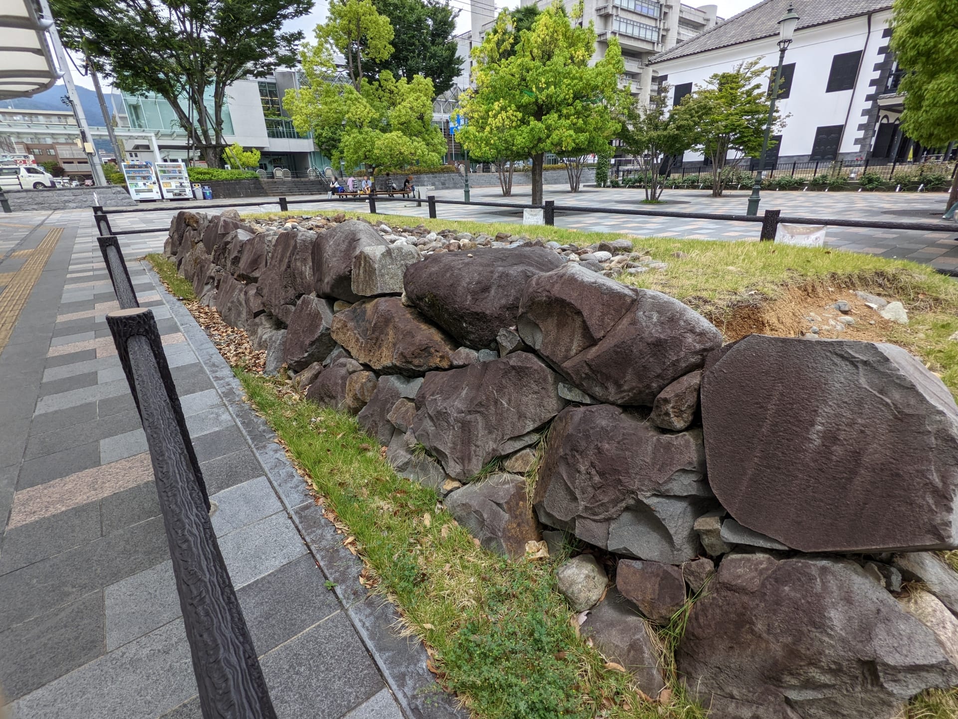 公園の舗装された歩道に沿って石積みの擁壁が並び、その背景には木々や建物が広がり、甲府の静かな雰囲気を醸し出しています。.