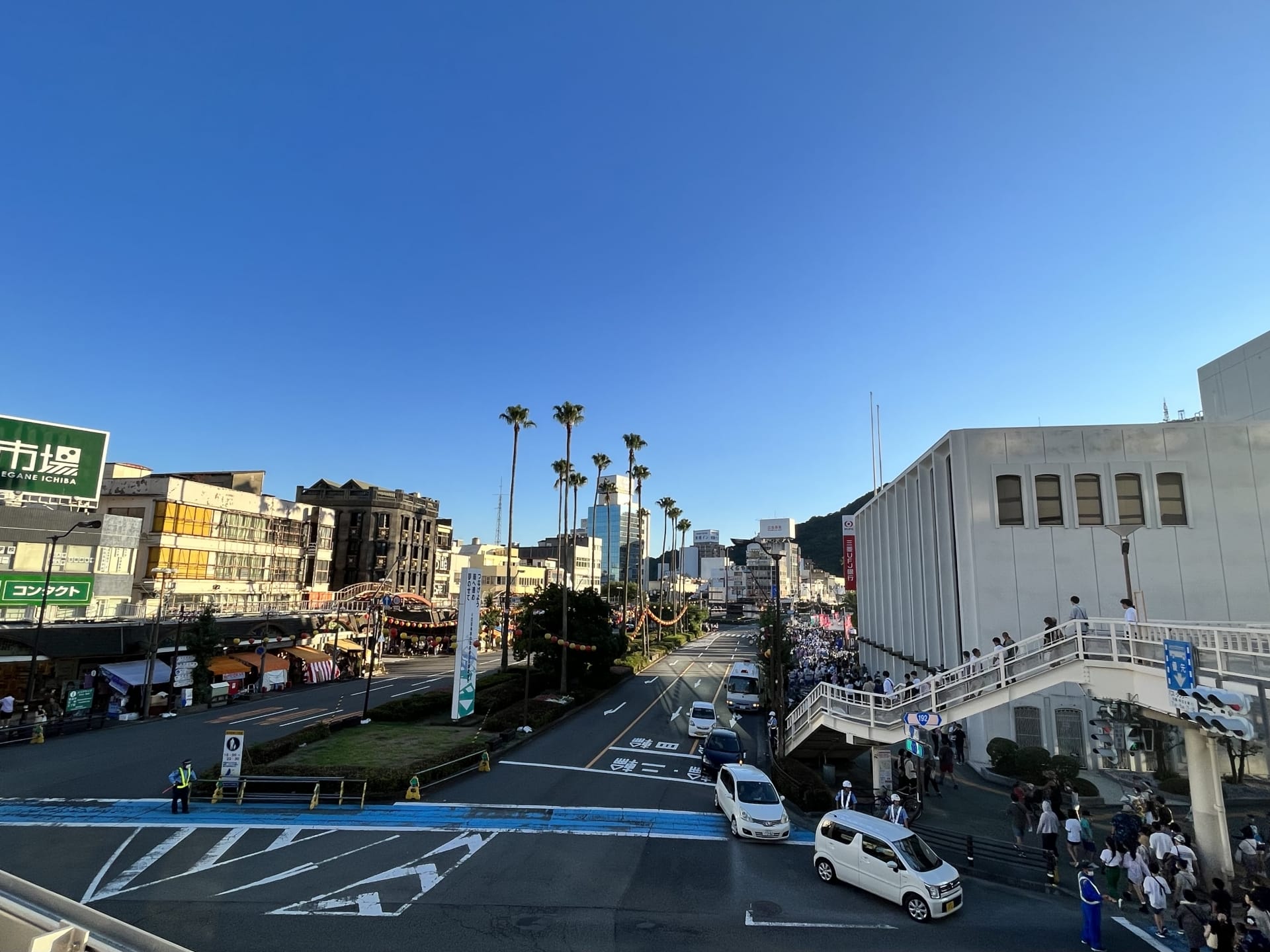 澄んだ青い空の下、建物やヤシの木が立ち並ぶ都会的な風景が広がります。通りは車と人で賑わっており、右側の歩道橋は活気に満ちた人々で賑わっており、文化がシームレスに交差する徳島、あるいはおそらく韓国語教室マンツーマンの近くの忙しい日々を思い出させます。.