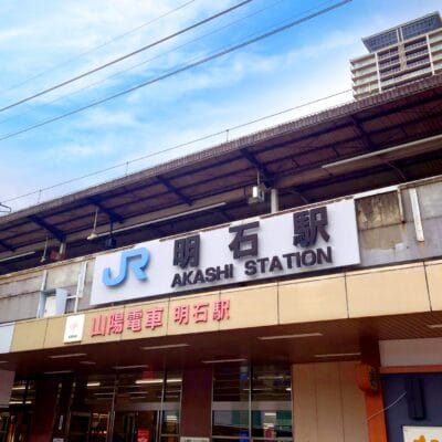 明石駅の正面図。JRのロゴと日本語、英語、韓国語の駅名標が掲げられています。建物は曇り空の下、明石の文化の融合を体現しています。.