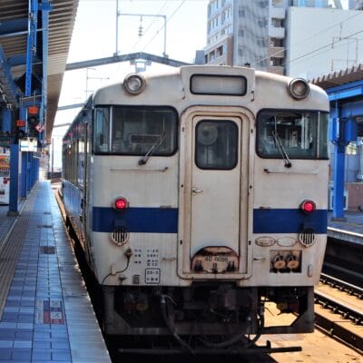 宮崎市の駅のホームで、都会のビル群を背景に電車を待つ電車。架線と青と白の洗練された外観が特徴。近くにある韓国語教室マンツーマンで個人に合わせた学習の機会を見つけて、旅行を充実させましょう。.