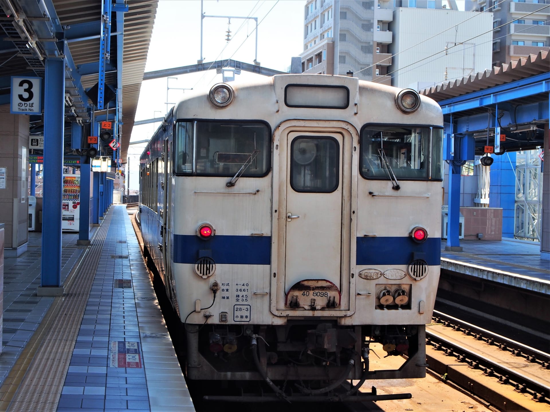 宮崎市の駅のホームで、都会のビル群を背景に電車を待つ電車。架線と青と白の洗練された外観が特徴。近くにある韓国語教室マンツーマンで個人に合わせた学習の機会を見つけて、旅行を充実させましょう。.