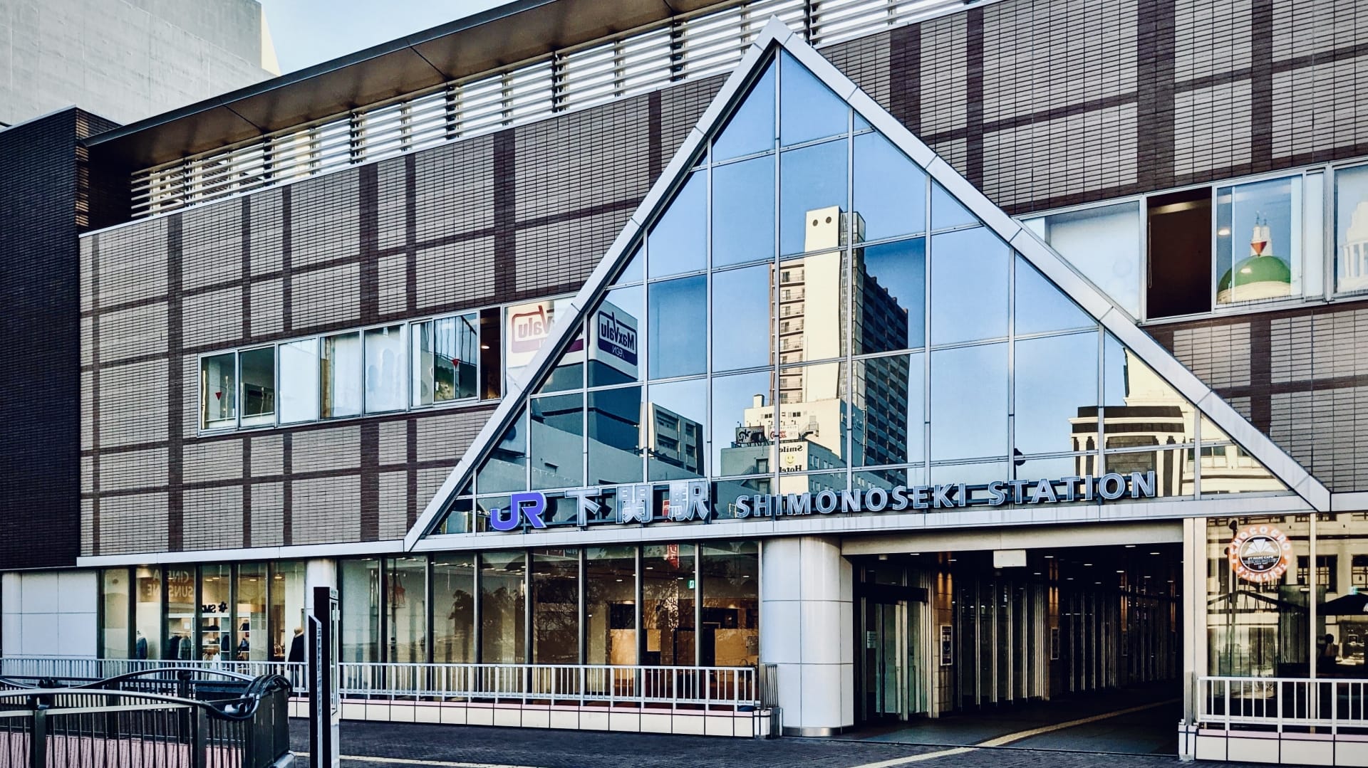 下関駅舎の正面図。三角ガラスのファサードと反射文字で駅名が表示され、窓に映る都市構造が特徴的。近くには、個人に合わせた語学レッスンを提供する韓国語教室マンツーマンがあります。.