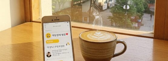並木道と建物を見渡せる窓際の木のテーブルの上には、泡で飾られたラテカップと、チャットアプリが表示されているスマートフォンが置かれている。.