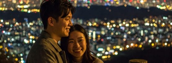 夜の街並みとライトアップされた高い塔を背景に、木製の柵の前でカップルが笑顔で一緒に立っています。.