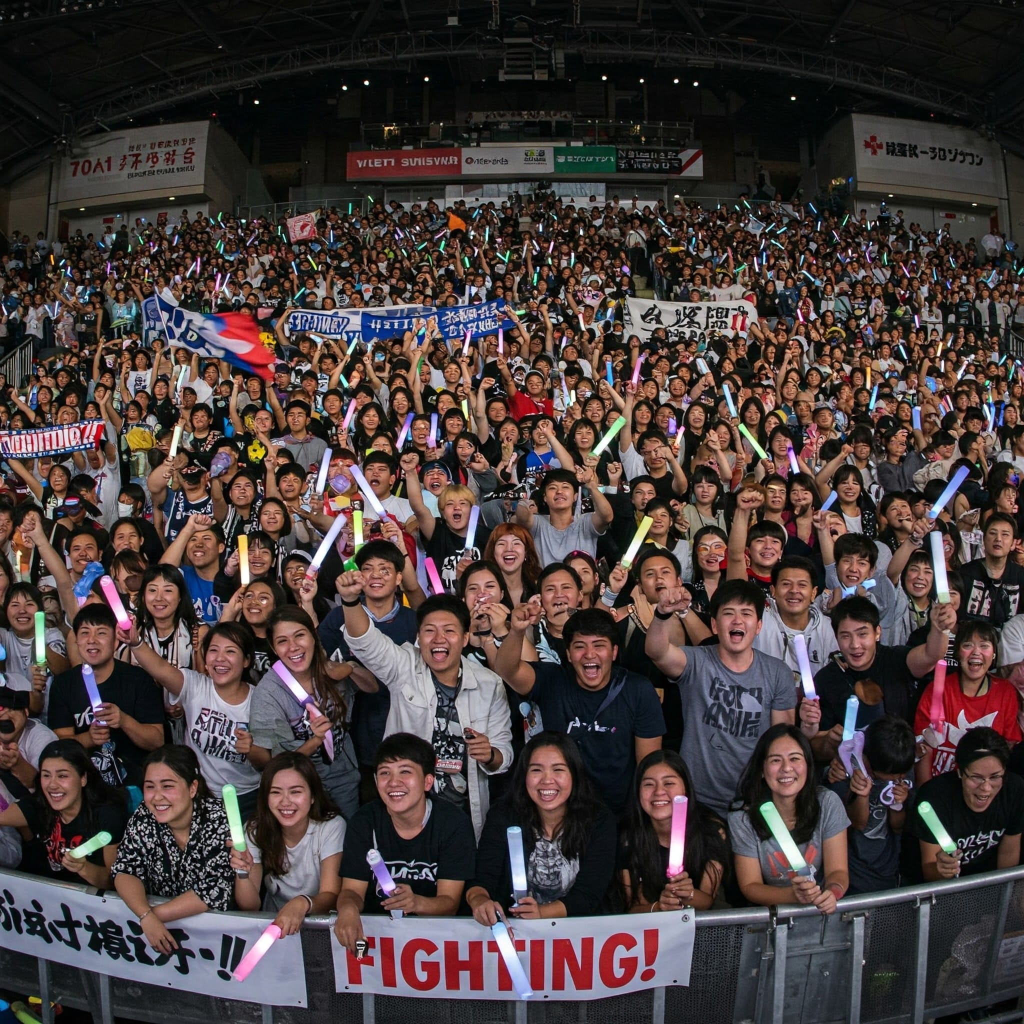 屋内アリーナに集まった大勢の興奮した観客は、色とりどりのライトスティックや横断幕を持ち、カメラに向かって熱狂的に笑顔を浮かべながら歓声を上げている。.