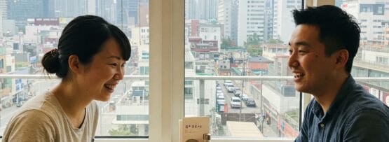 2 人がカフェのテーブルに座り、目の前にコーヒー カップとメニューを置いて笑顔で話しています。背後の大きな窓からは街のスカイラインが見えます。.