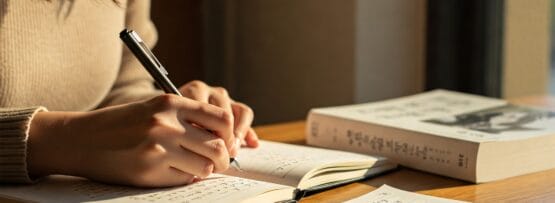 木製のテーブルの上に、近くの本を開いてノートに書いている人がいます。日光がページに影を落としています。.