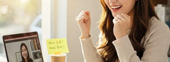 女性が机に座り、ノートパソコンでビデオ通話をしながら、笑顔で拳を突き上げ、興奮している。彼女の前には、書類、本、付箋が広げられている。.
