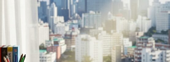 開いたプランナー、積み重ねられた本、ホルダーに立てられたペン、コーヒーマグが置かれた木製の机が、近代的な街のスカイラインを見渡す窓のそばに置かれています。.