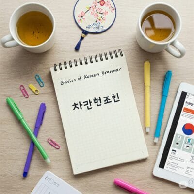 「韓国語の文法の基礎」と題された韓国語のテキストが書かれたメモ帳。周りにはティーカップ、ペン、蛍光ペン、ペーパークリップ、扇風機、韓国語のウェブサイトが表示されているタブレットが置かれている。.