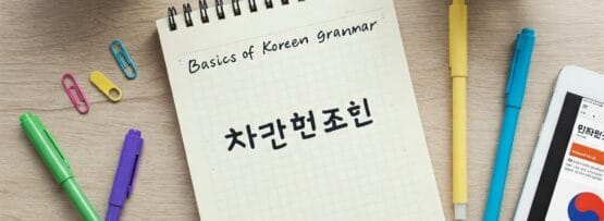 「韓国語の文法の基礎」と題された韓国語のテキストが書かれたメモ帳。周りにはティーカップ、ペン、蛍光ペン、ペーパークリップ、扇風機、韓国語のウェブサイトが表示されているタブレットが置かれている。.