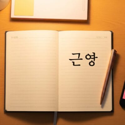 木製の机の上に、韓国語の文字が書かれた開いたノート、ペン、本、韓国ドラマが表示されているスマートフォン、コーヒーカップ、タブレットが置いてあります。.