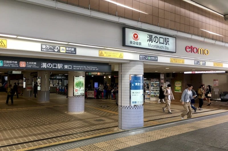 日本の溝の口駅の入り口。中に複数の看板、歩く人々、自動販売機、店が見える。.