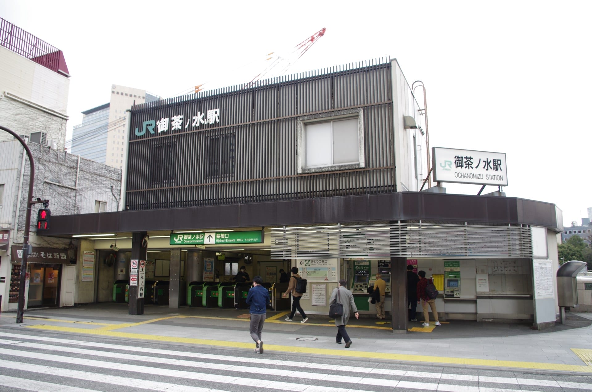東京・御茶ノ水駅の入り口。灰色の長方形の建物の下にある改札口から人々が出入りしている。.