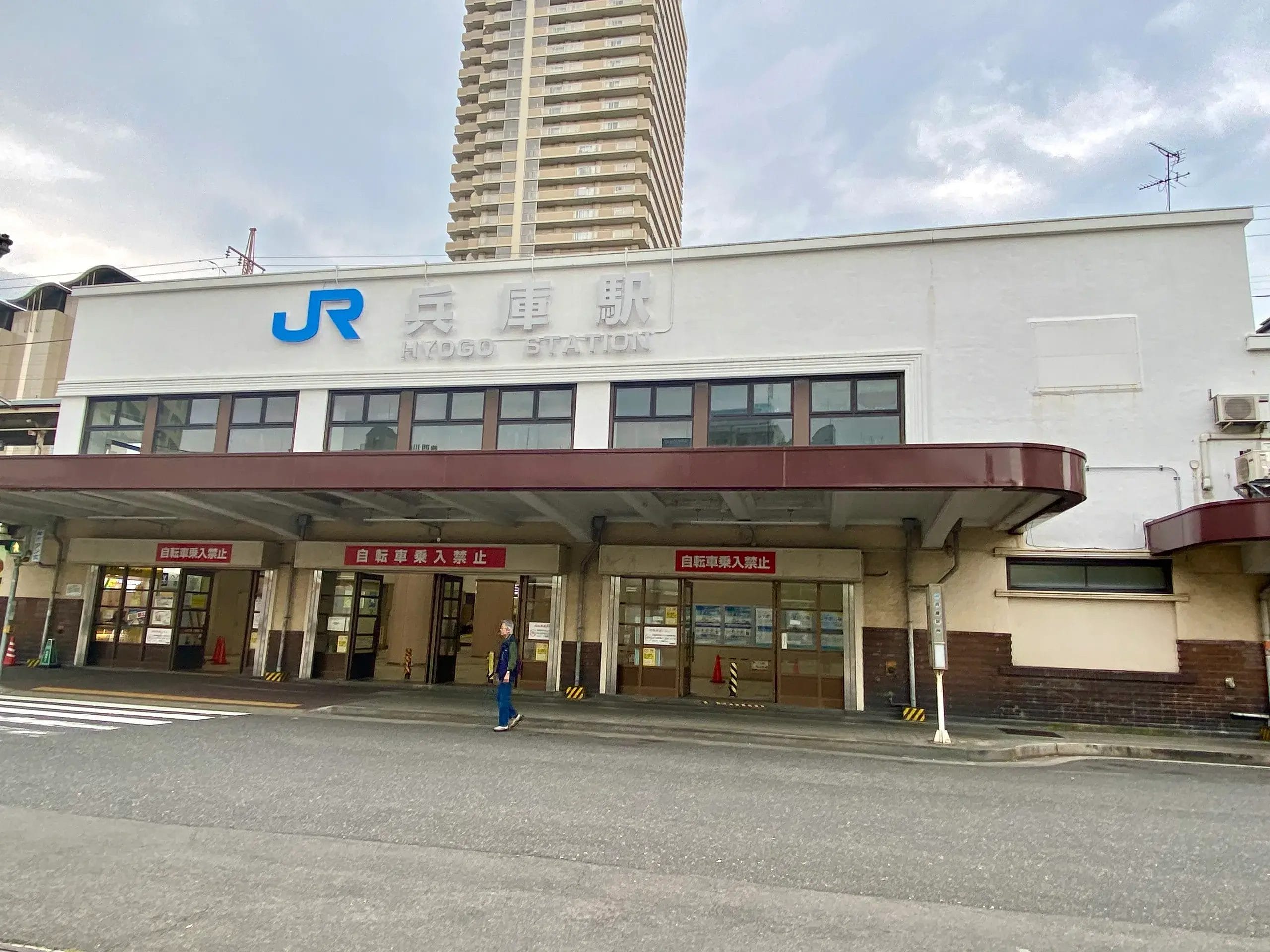 JRのロゴ、ガラス窓、赤い日よけのある共同駅舎。正面を人が横切り、背景に高いビルが見える。.