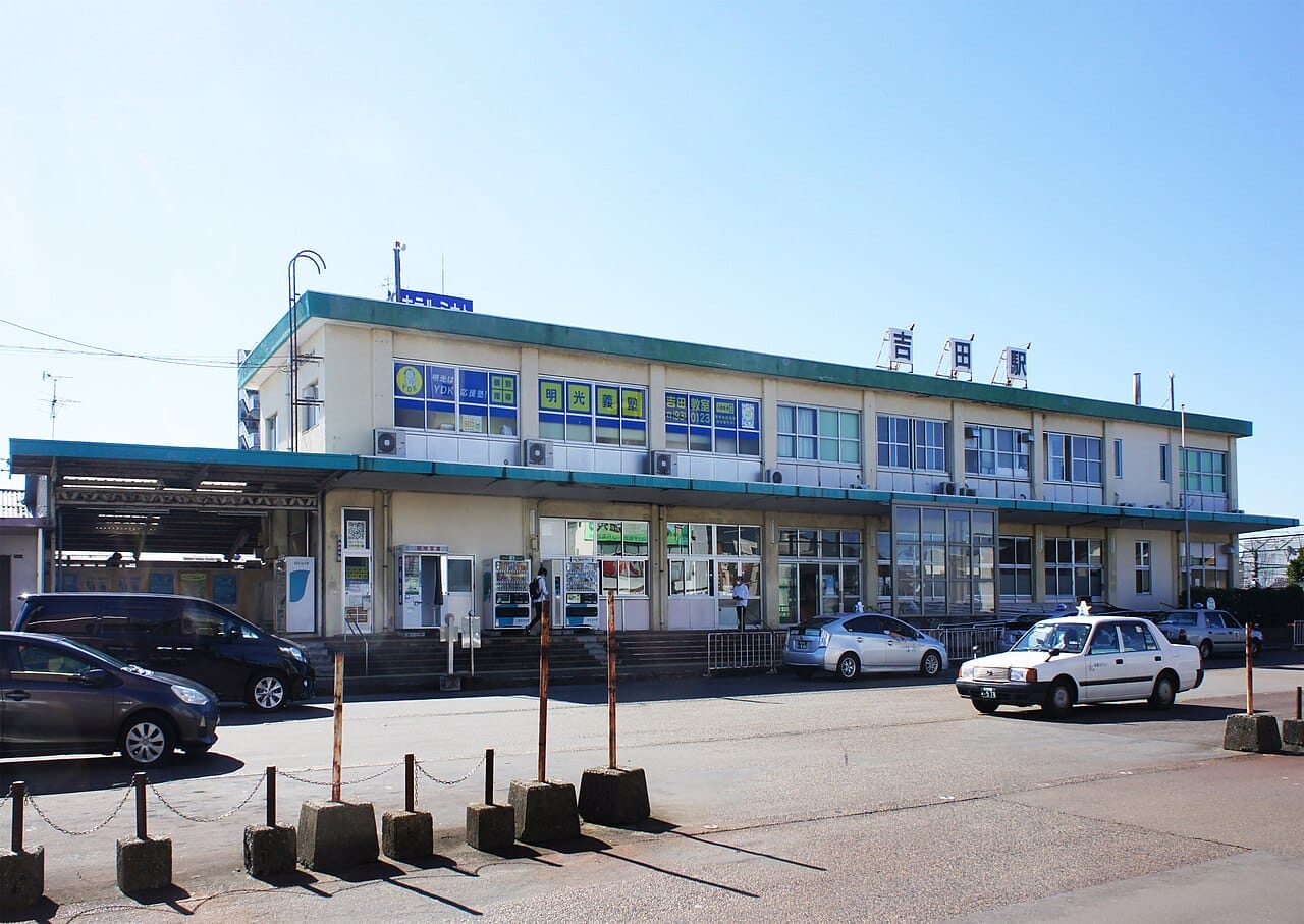 晴れ渡った青空の下、2階建ての駅舎にはいくつもの窓と看板があり、正面には車やタクシーが停まっている-吉田の韓国語教室の口コミで話題のスポットです。.