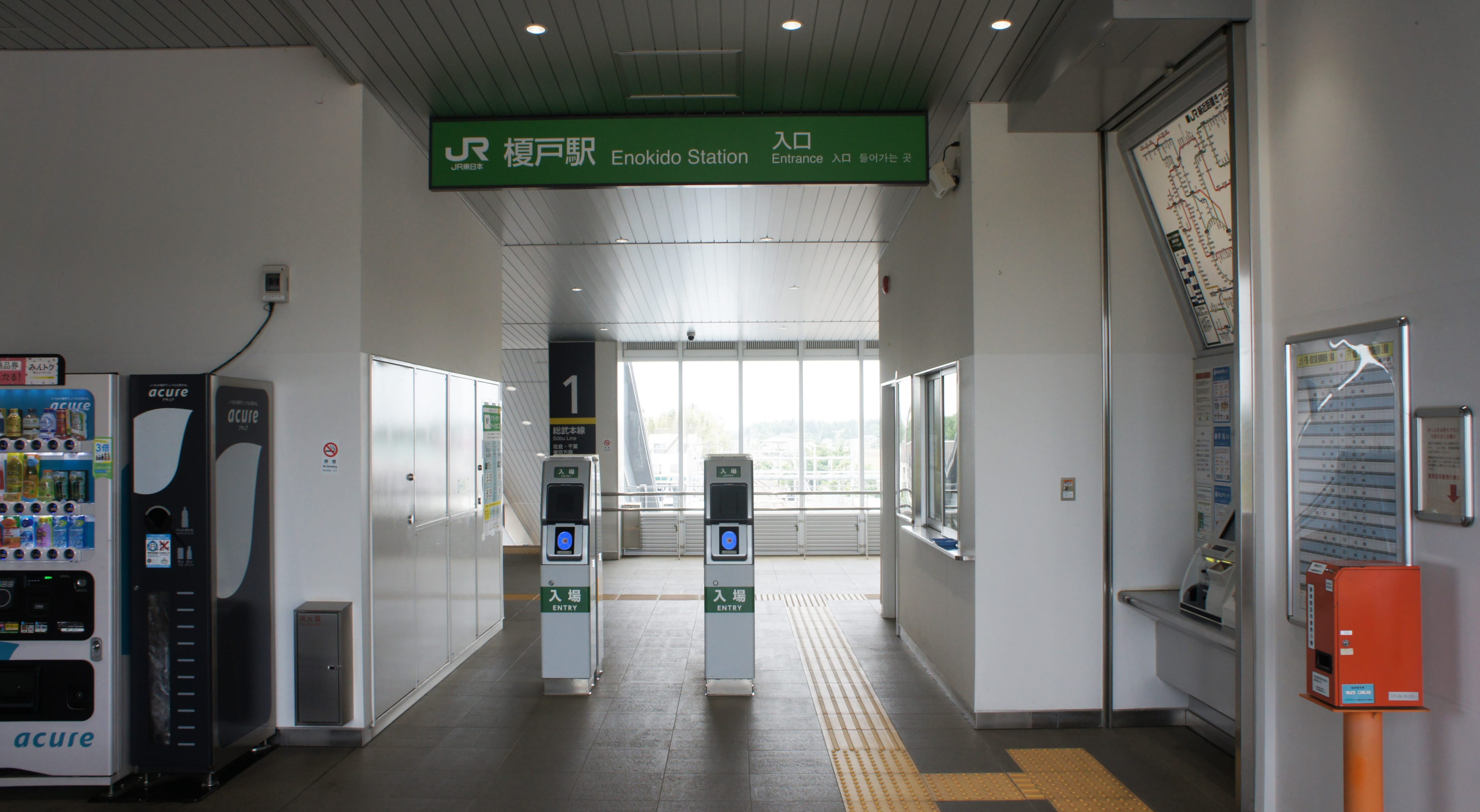 改札口、自動販売機、切符販売機、公衆電話、そして頭上に日本語と英語の駅名標がある榎戸駅の入口付近。.