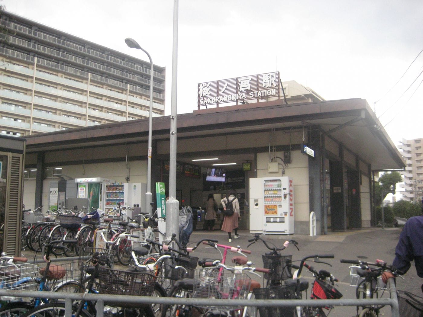桜ノ宮駅の入り口の外に駐輪された自転車。いくつかの自動販売機が見え、人が入ってくる。.